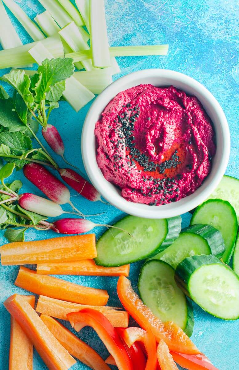 A bright blue table setting with a tub of beetroot hummus and lots of fresh crunchy vegetables surrounding including sliced cucumber, peppers, carrots, celery and radish.
