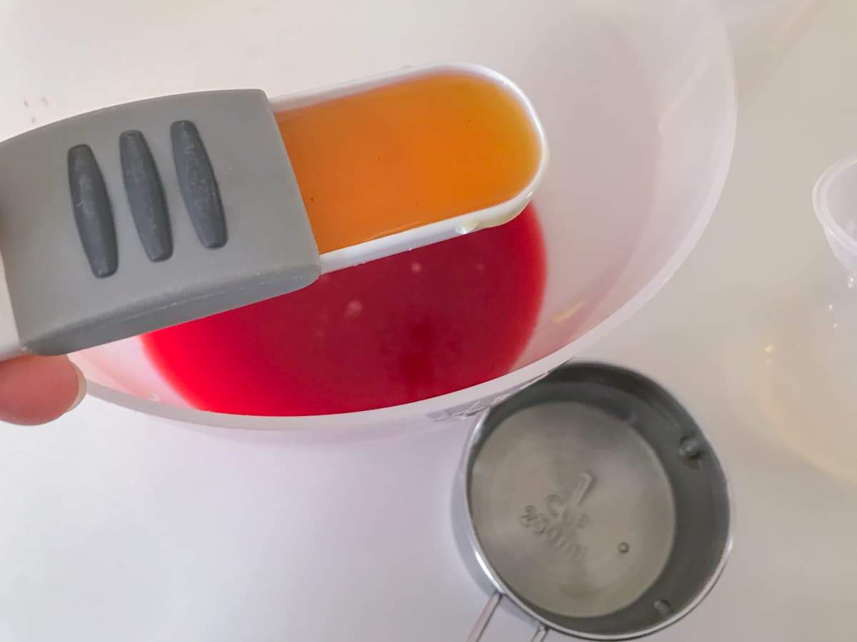 A spoon of golden syrup being added to a jug of fresh blood orange juice and a measuring cup of sugar syrup.