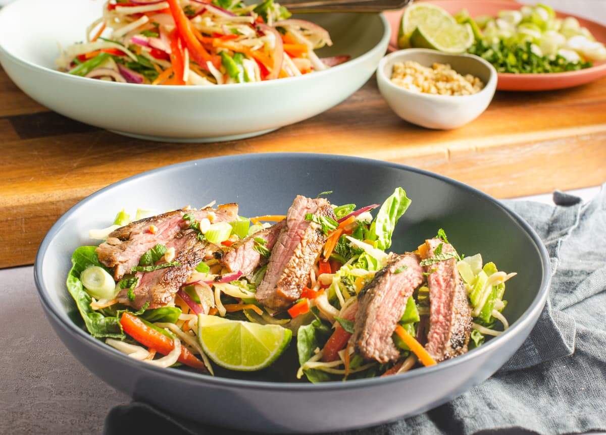 A table setting of a grey bowl with sliced marinated asian beef salad on a bed of colourful slaw.