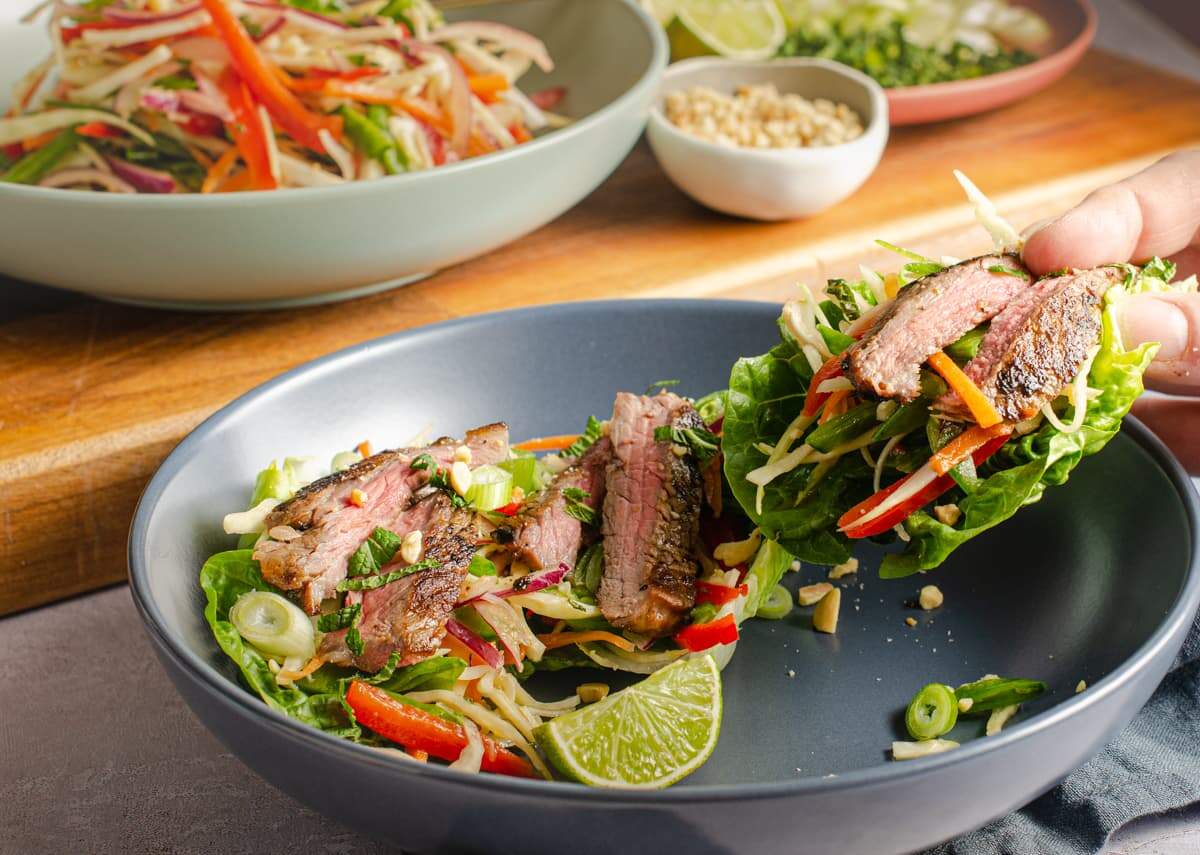 Slices of Asian marinated beef on a colourful slaw base with a wedge of lime showing someone lifting the food up to eat.