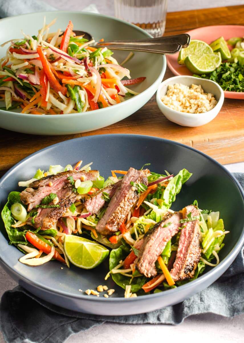 A table setting of a grey bowl with sliced marinated asian beef salad on a bed of colourful slaw a wooden board to the back with a bowl of slaw, peanuts and sliced lime and chopped corriander.