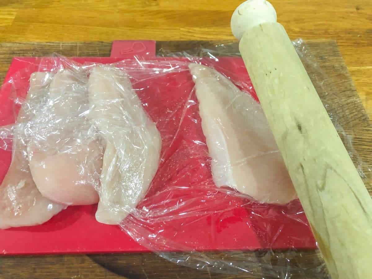 Chicken breast on a red chopping board covered in cling film and a rolling pin next to it.