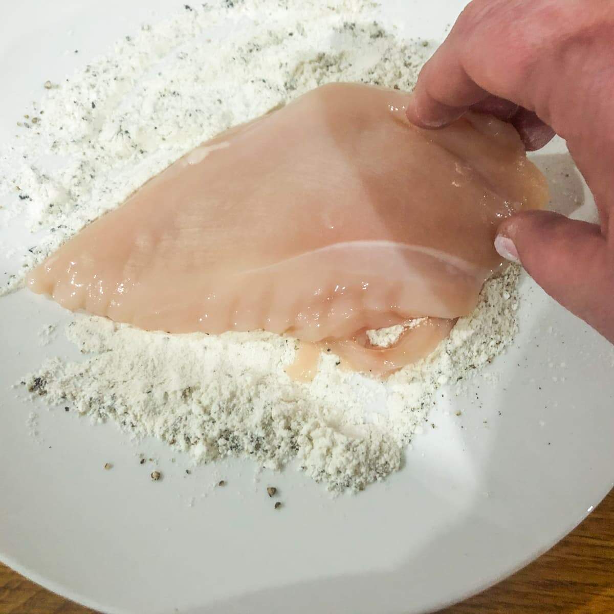 Chicken breast being tossed in seasoned flour in a white bowl on a wooden board.