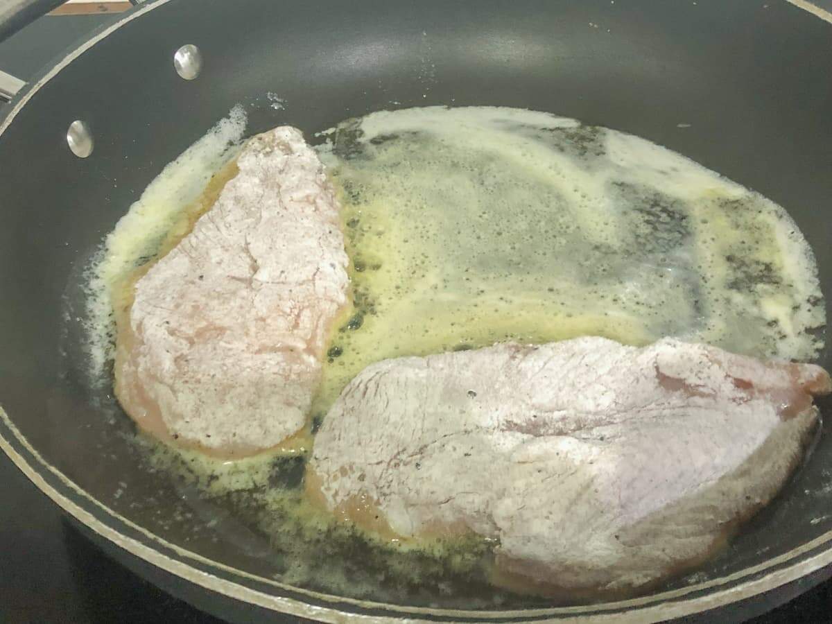 two chicken breast coated in flour being pan fried in a black frying pan with butter.