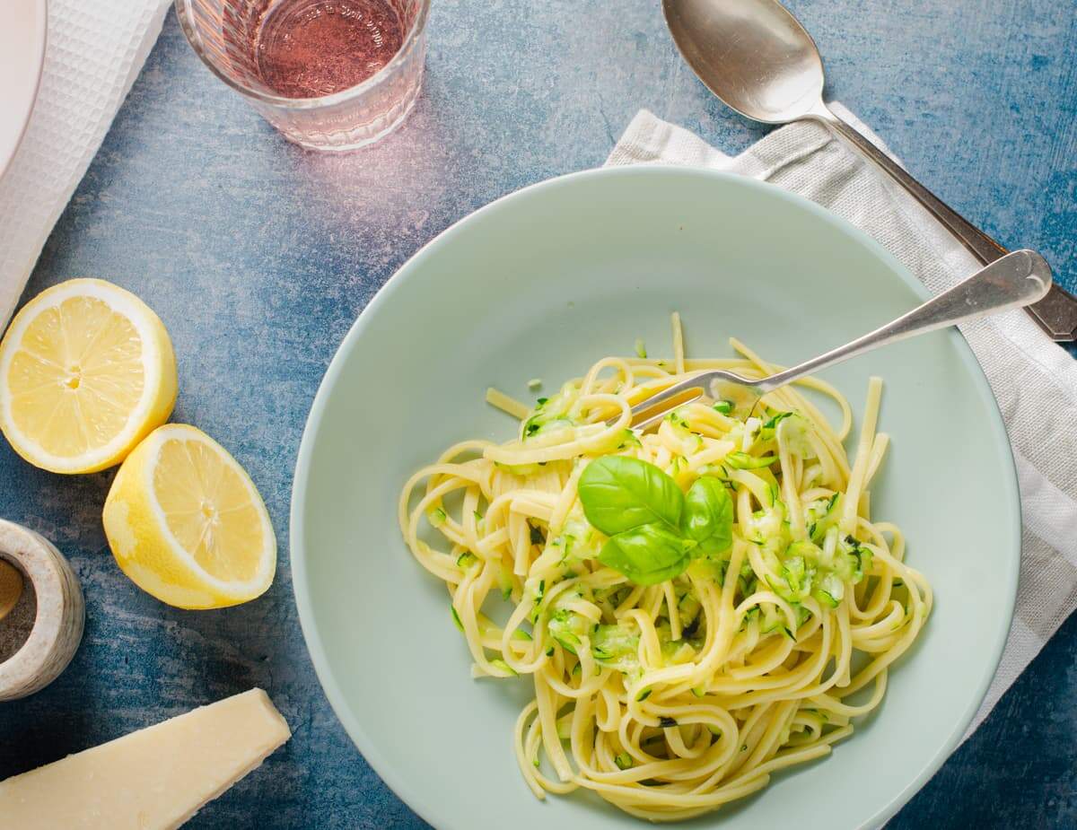 A table setting of a fresh bowl of linguine with courgette and lemon with a fork in and to the side a lemon cut in half, a block of parmesan cheese and come salt with a glass of rose wine.