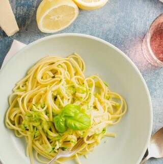 A top down view of a pale green bowl containing freshly cooked linguine with courgette some lemons to the back with parmesan cheese and a glass of wine.