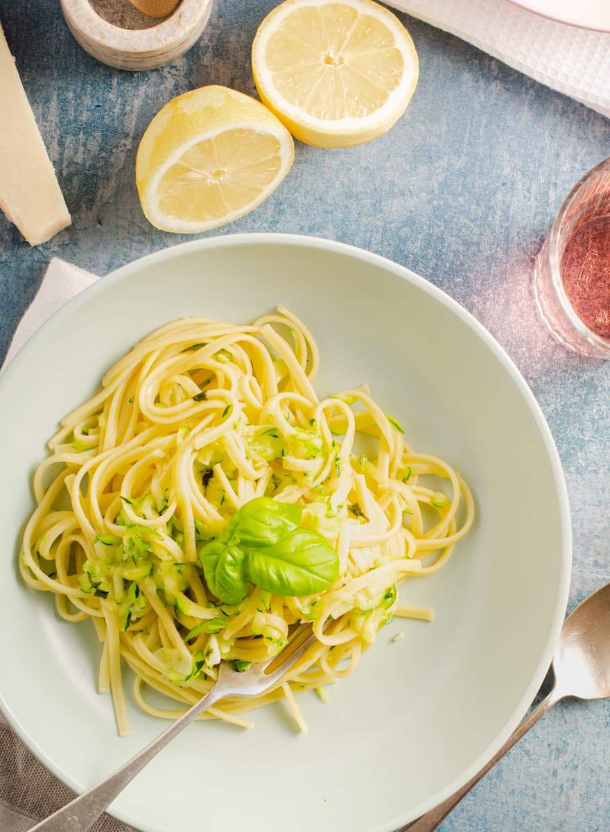 A top down view of a pale green bowl containing freshly cooked linguine with courgette some lemons to the back with parmesan cheese and a glass of wine.