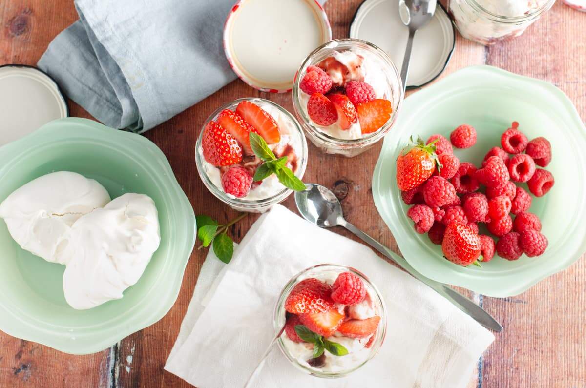 A picnic table setting of green bowls filled with meringues, fresh raspberries and strawberries and some linen napkins and 4 glass jam jars filled with Eton Mess and topped with fresh berries and mint.