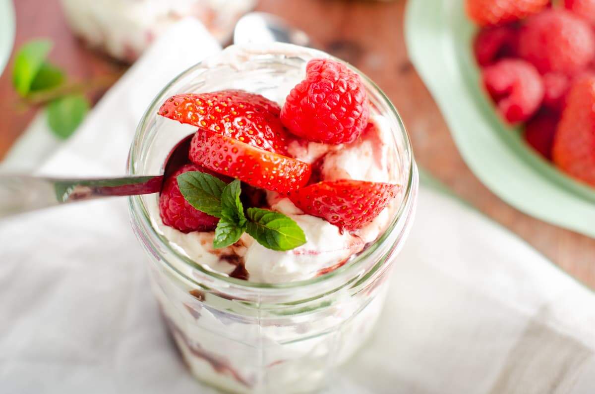 A close up of a glass jam jar filled with Eton Mess and topped with fresh raspberries and strawberries and a spoon in the jar sitting on a white linen napkin and a bowl of fresh berries to the back