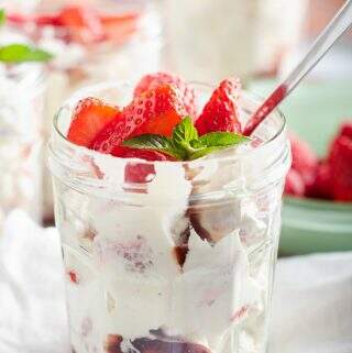 A single jam jar filled with Eton Mess topped with more fresh berries and a sprig of mint with more jars in the background and a green bowl of fresh berries to the side.