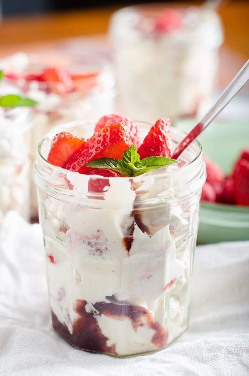 A single jam jar filled with Eton Mess topped with more fresh berries and a sprig of mint with more jars in the background and a green bowl of fresh berries to the side.