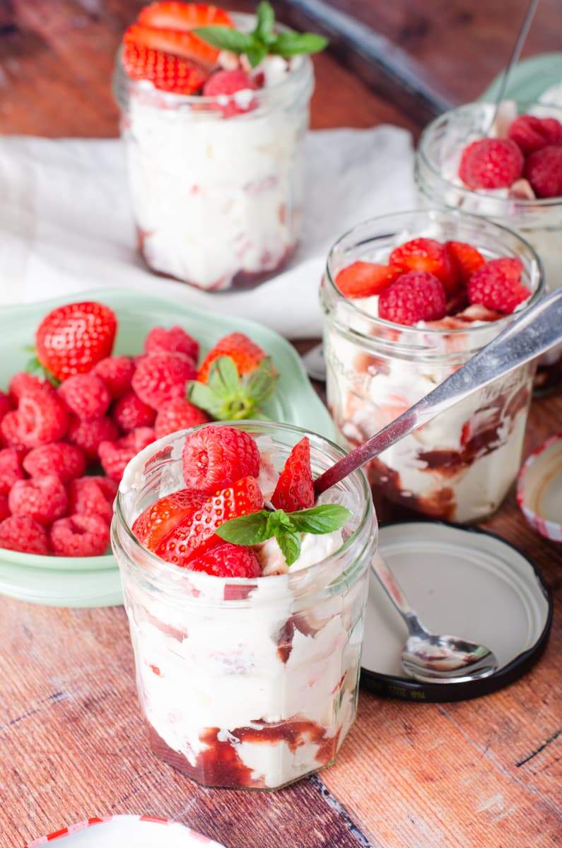 4 jam jars filled with a strawberry and raspberry eton mess, topped with more fresh berries, some with spoons and the jar lids on the table, all ready for a picnic