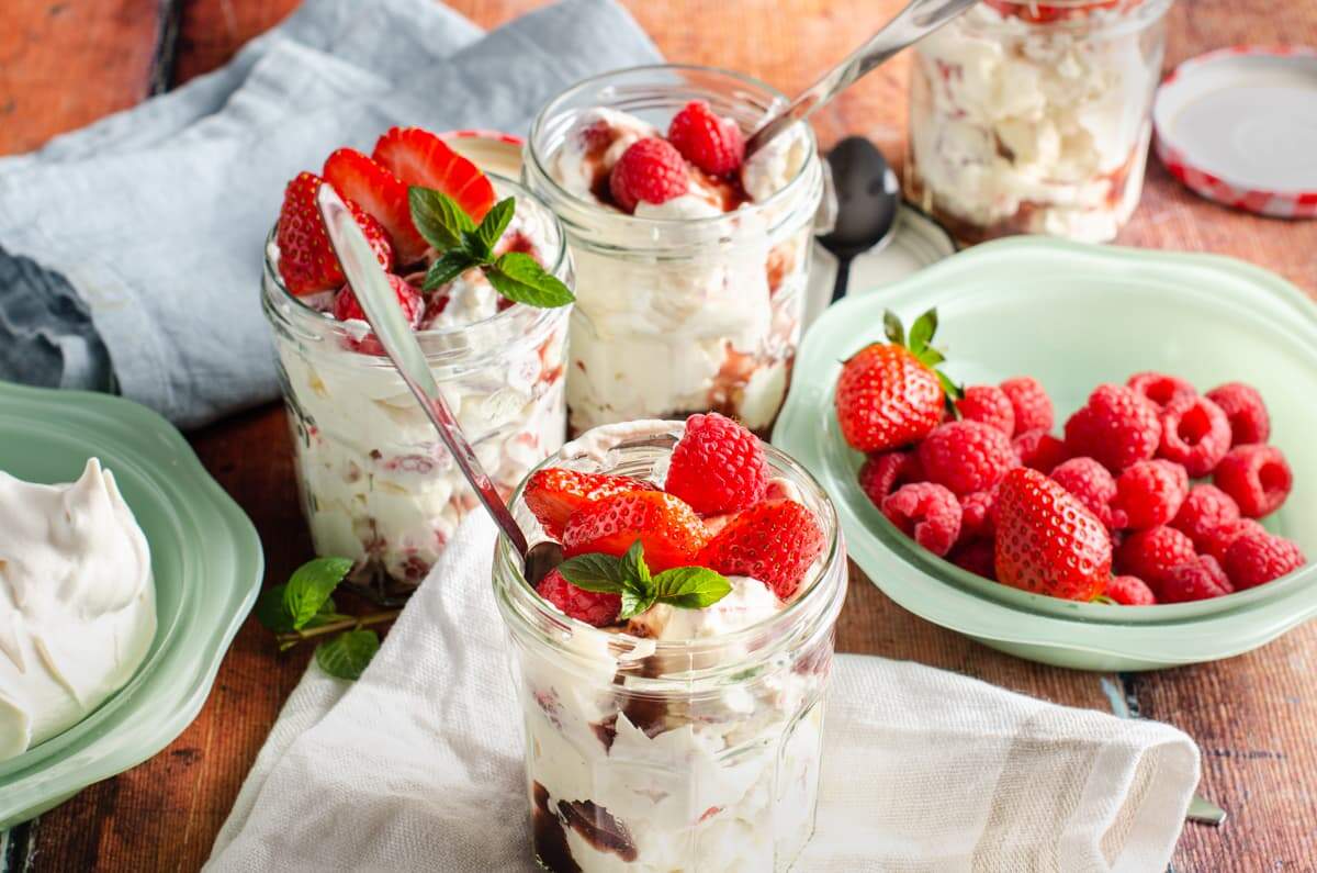 A picnic table setting of green bowls filled with meringues, fresh raspberries and strawberries and some linen napkins and 4 glass jam jars filled with Eton Mess and topped with fresh berries and mint.