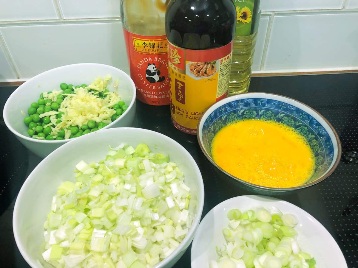Ingredients laid out for egg fried rice including diced onions, spring onions, eggs, peas, garlic, ginger, soy sauce, oyster sauce and oil