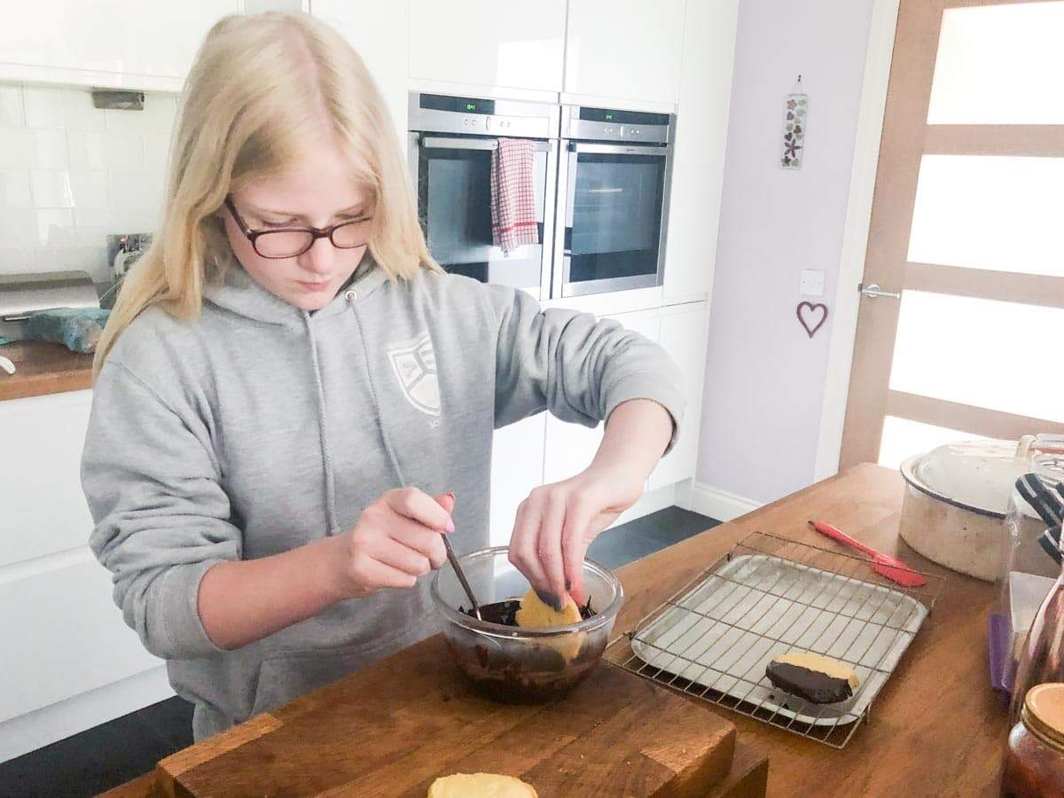 Young girl in a kitchen dipping orange biscuits in melted chocolate.