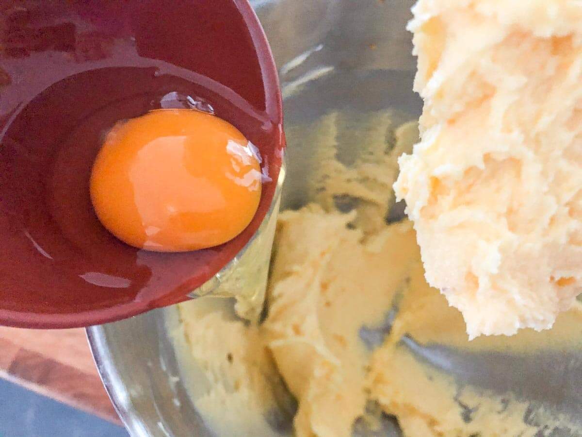 Creamed butter and sugar in a bowl of a stand mixer and an egg being added.