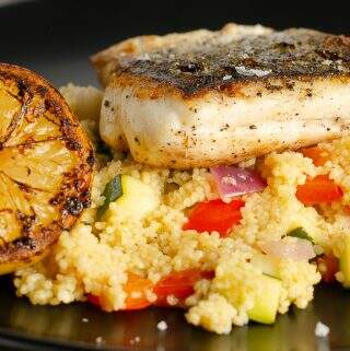 A closeup view of a dish of pan fried hake on a bed of lemon, pepper and courgette couscous with a grilled lemon on the side on a black plate.