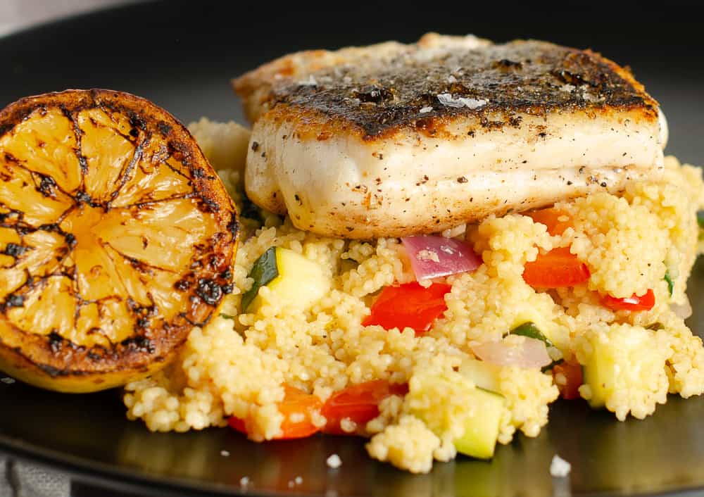 A closeup view of a dish of pan fried hake on a bed of lemon, pepper and courgette couscous with a grilled lemon on the side on a black plate.