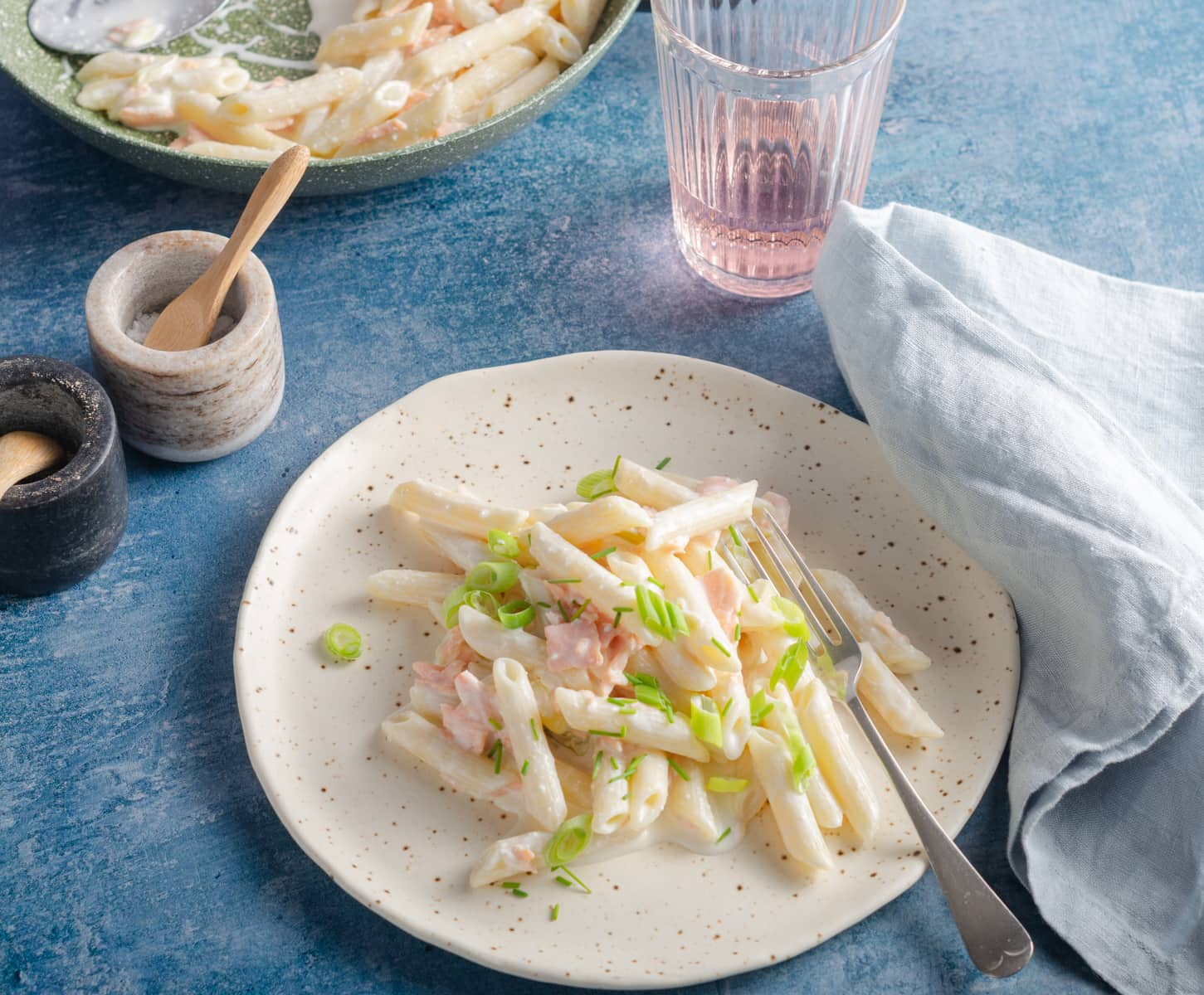 Smoked salmon pasta topped with chives and spring onion on a blue table and a pan to the back holding the rest of the dish