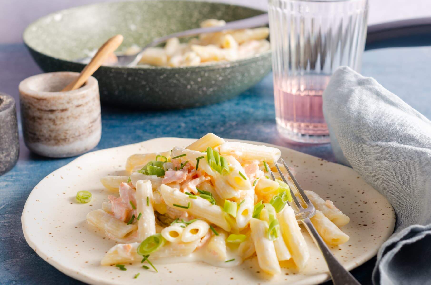 A table setting of smoked salmon penne and a glass of pink juice to the back and a frying pan with the rest of the dish.