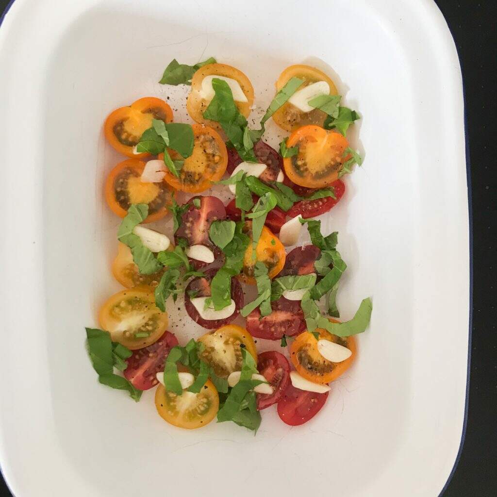 red, yellow and orange cherry tomatoes halved and topped with sliced garlic, oil, salt and pepper and fresh herbs in a white roasting tray.