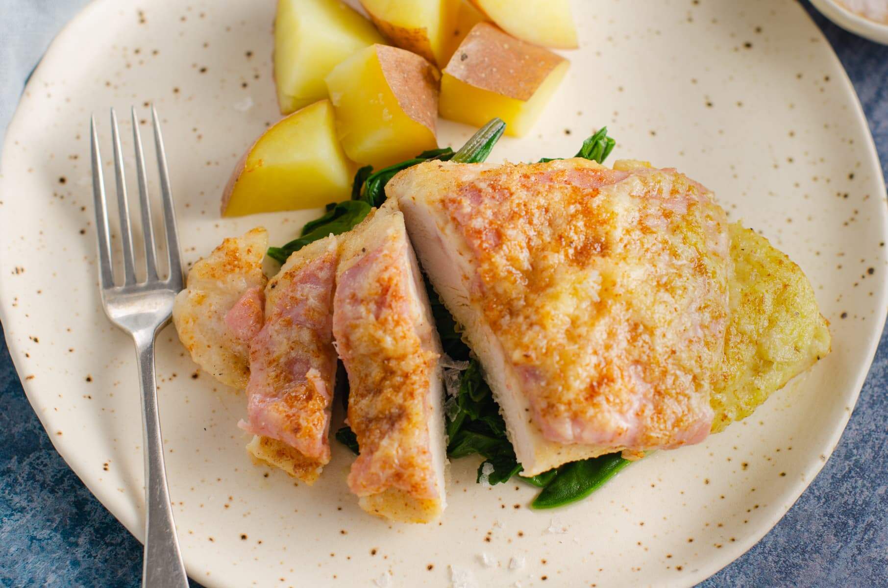 A plate of pan fried chicken topped with ham and parmesan cheese served with new potatoes and wilted greens.