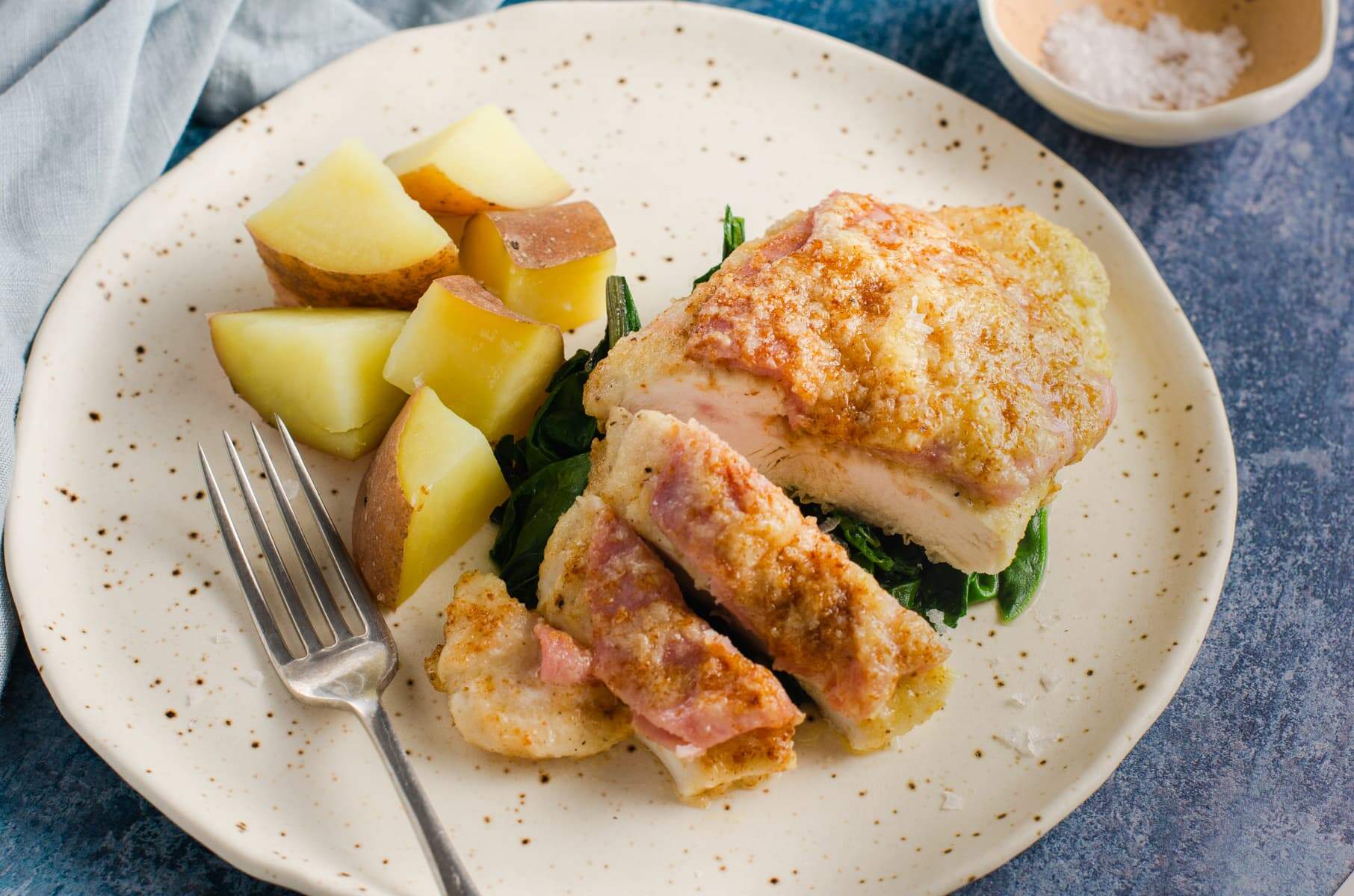 A ceramic speckled plate with a serving of pan fried chicken breast topped with sliced ham and parmesan cheese on a bed of wilted greens and served with boiled cubed potatoes.