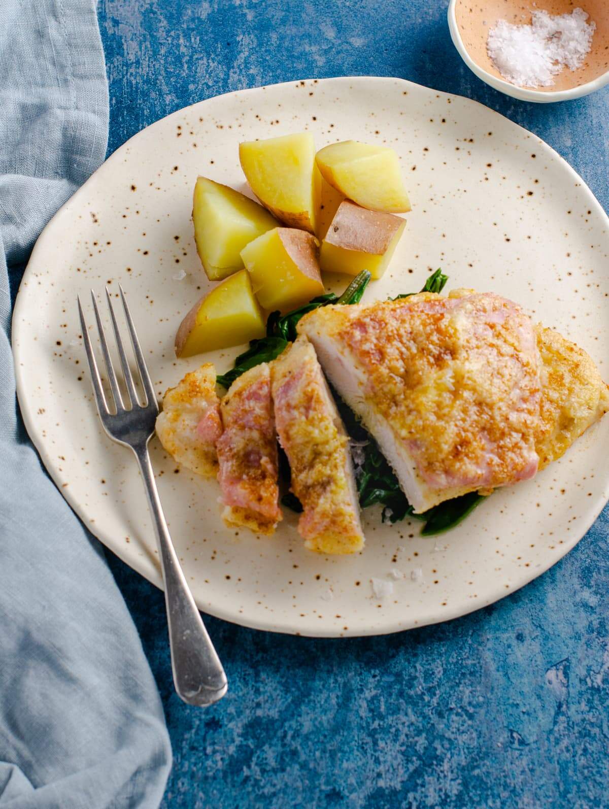 A speckled ceramic plate of sauteed chicken with ham and parmesan cheese served with boiled boiled and pan fried greens on a blue background with a pale blue napkin.