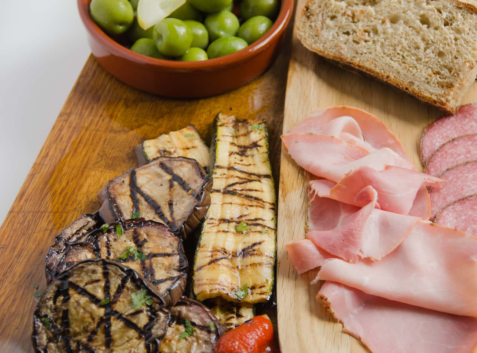 An antipasti board of marinated aubergine, courgette and peppers with cured meats, bread and olives.