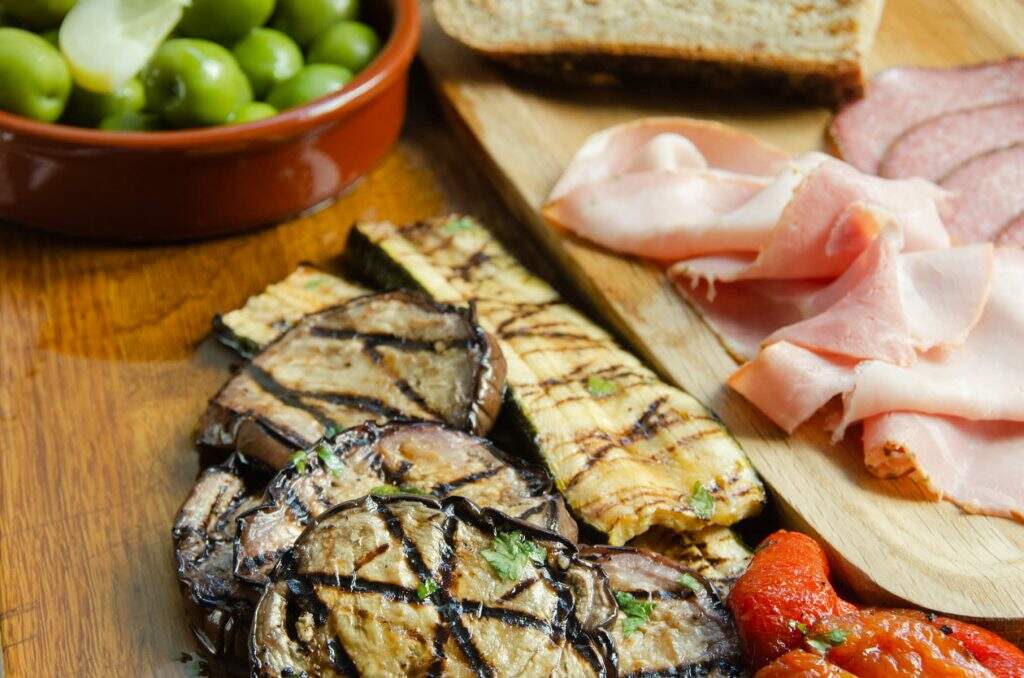 A wooden platter containing chargrilled aubergine, courgettes and peppers, alongside some sliced ham and salami and a bowl of green olives.