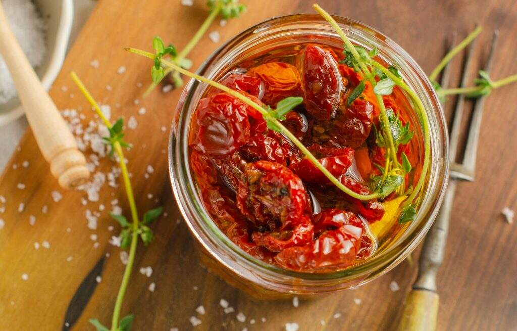 A jar of oven roasted tomatoes in oil with fresh thyme sitting on a wooden board sprinkled with sea salt and an old fork to the side.