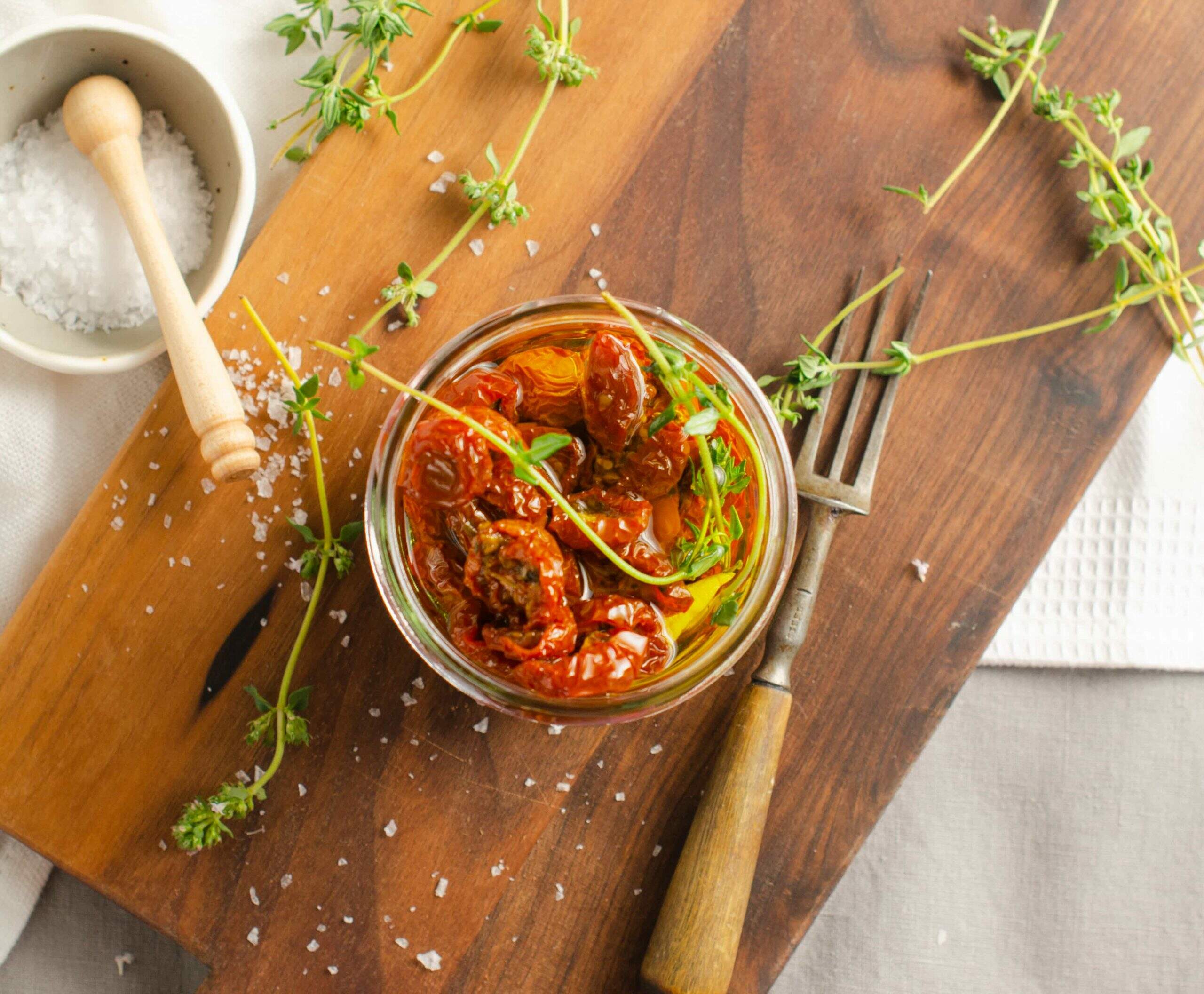 A jar filled with oven roasted tomatoes in oil with thyme and sitting on a dark wooden board, sea salt sprinkled and in a pinch bowl and a wooden handled fork for serving.
