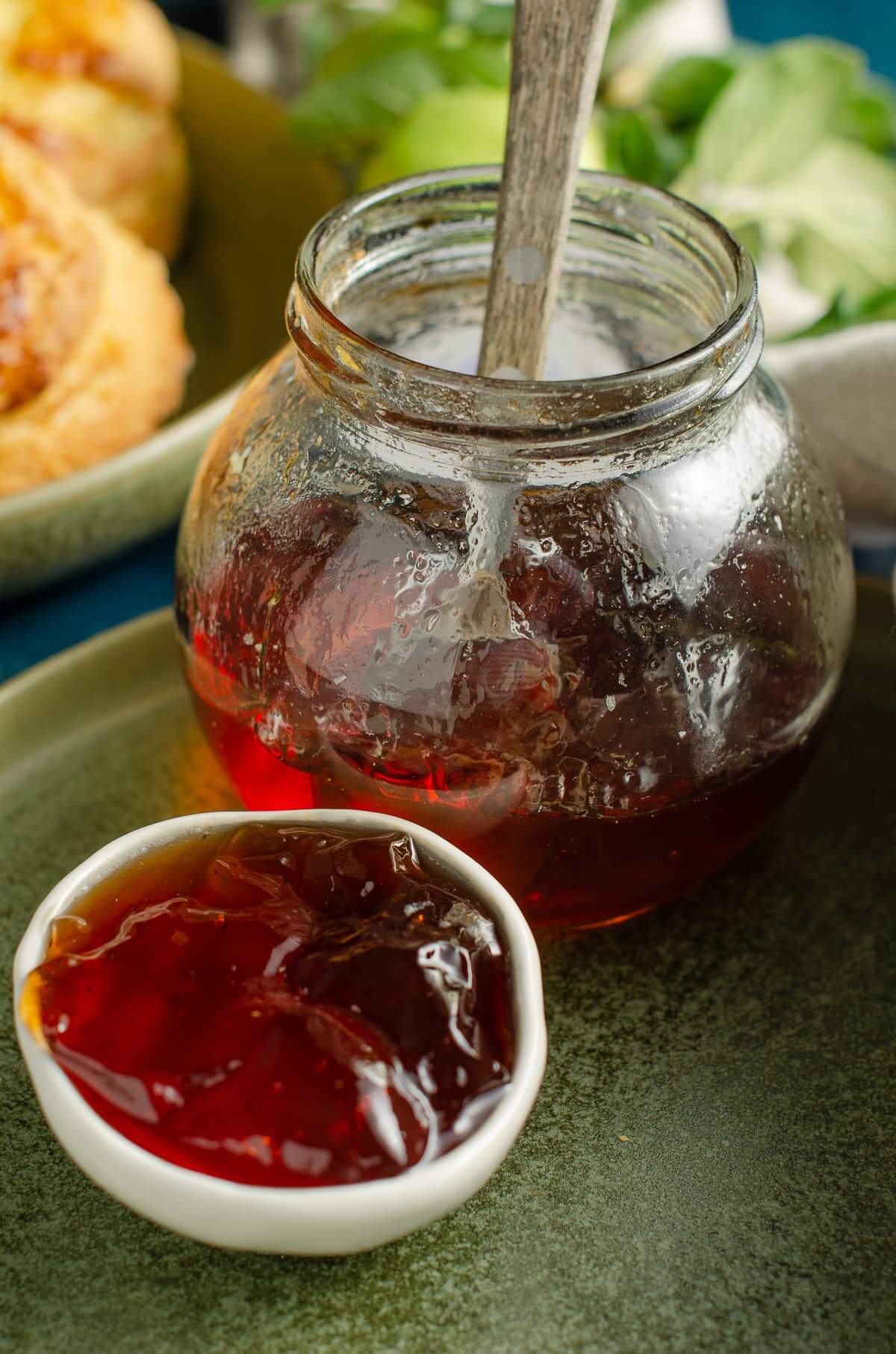 A round bellied jar of apple jelly jam half full with a spoon inside and a small serving bowl of jam in front.