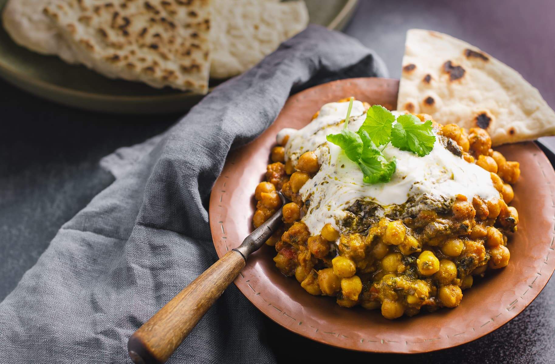 Chickpea curry topped with spiced yogurt in a copper bowl and served with fresh flatbreads and a blue denim shaded napkin.