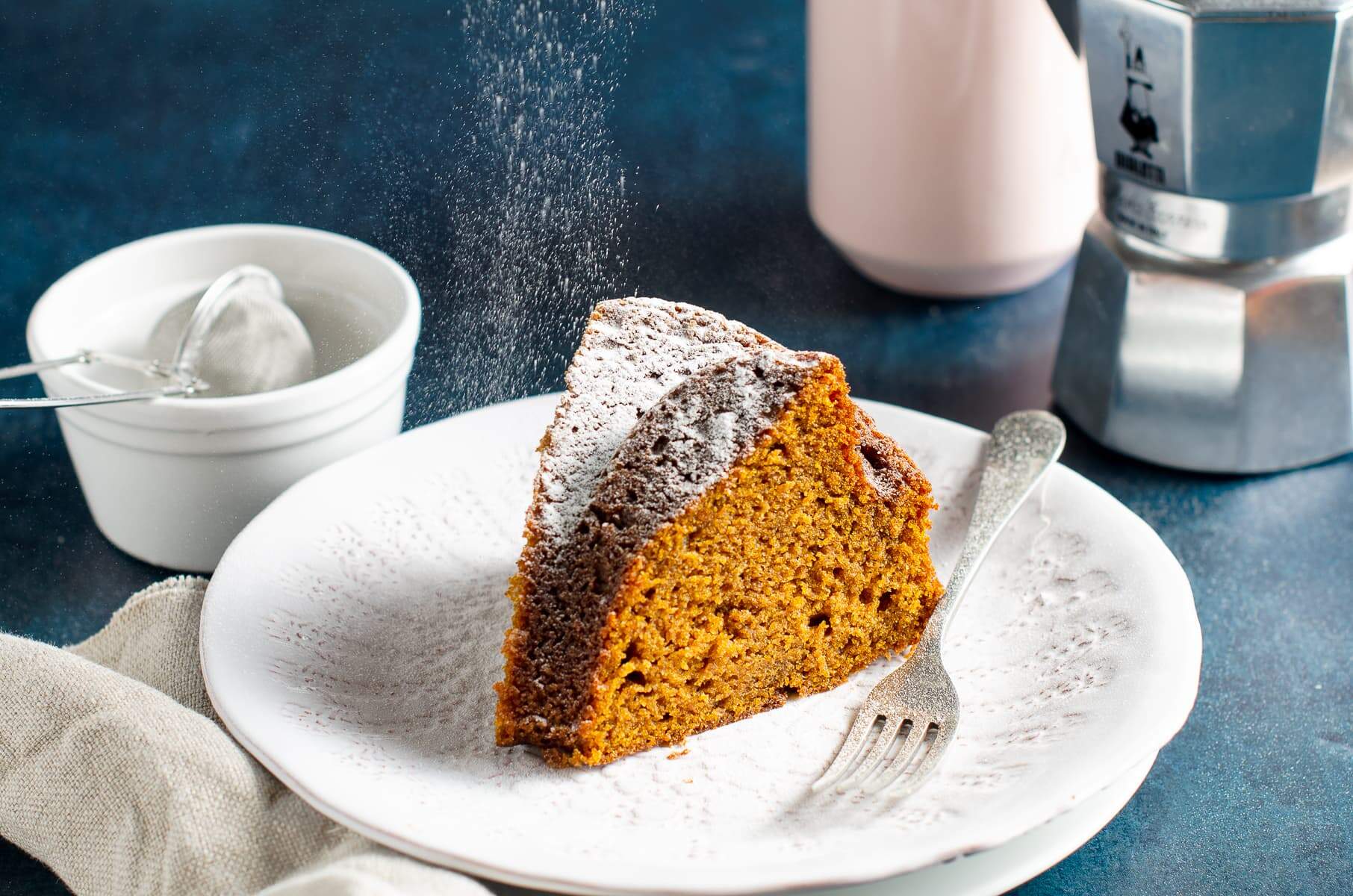 A slice of pumpkin cake with sugar being dusted over the top served with a cup of coffee.