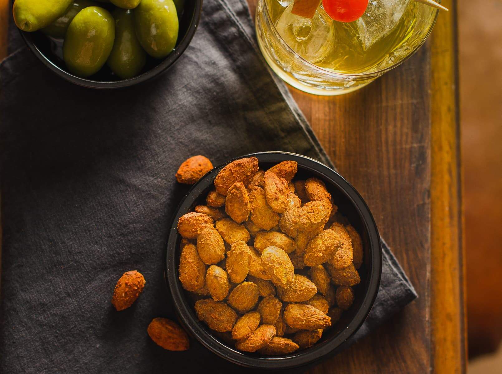 A black tub of spiced nuts served with a whisky cocktail and olives on a mahogany table end.