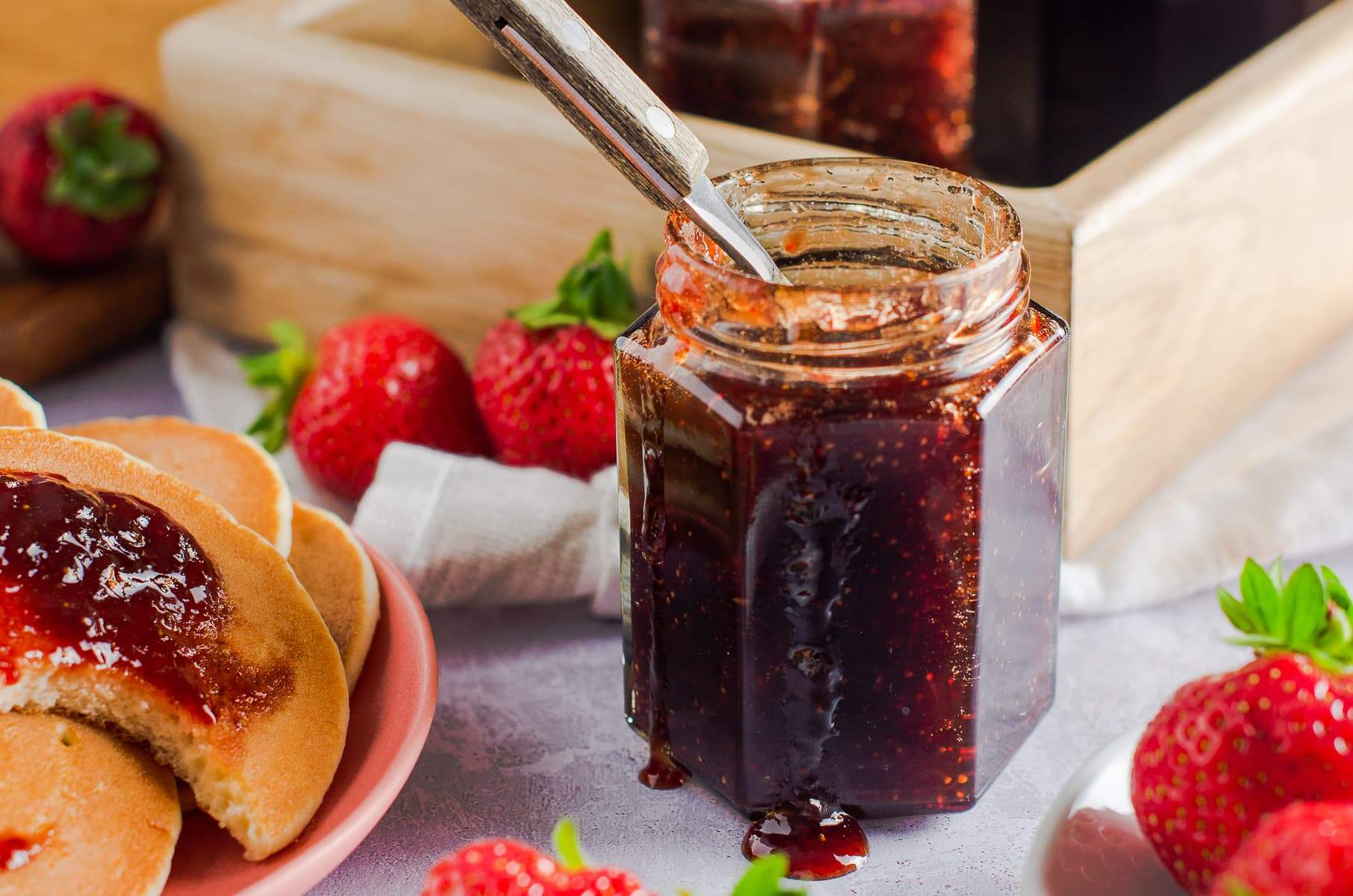 A jar of jam, pancakes topped with strawberry jam, some fresh berries and extra jars to the back.
