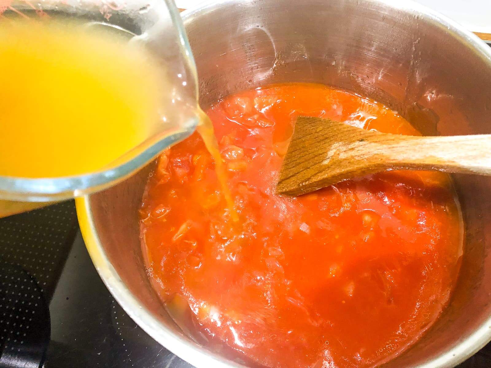 A hand pouring vegetable stock to a pan of simmering onions and tomato juice.