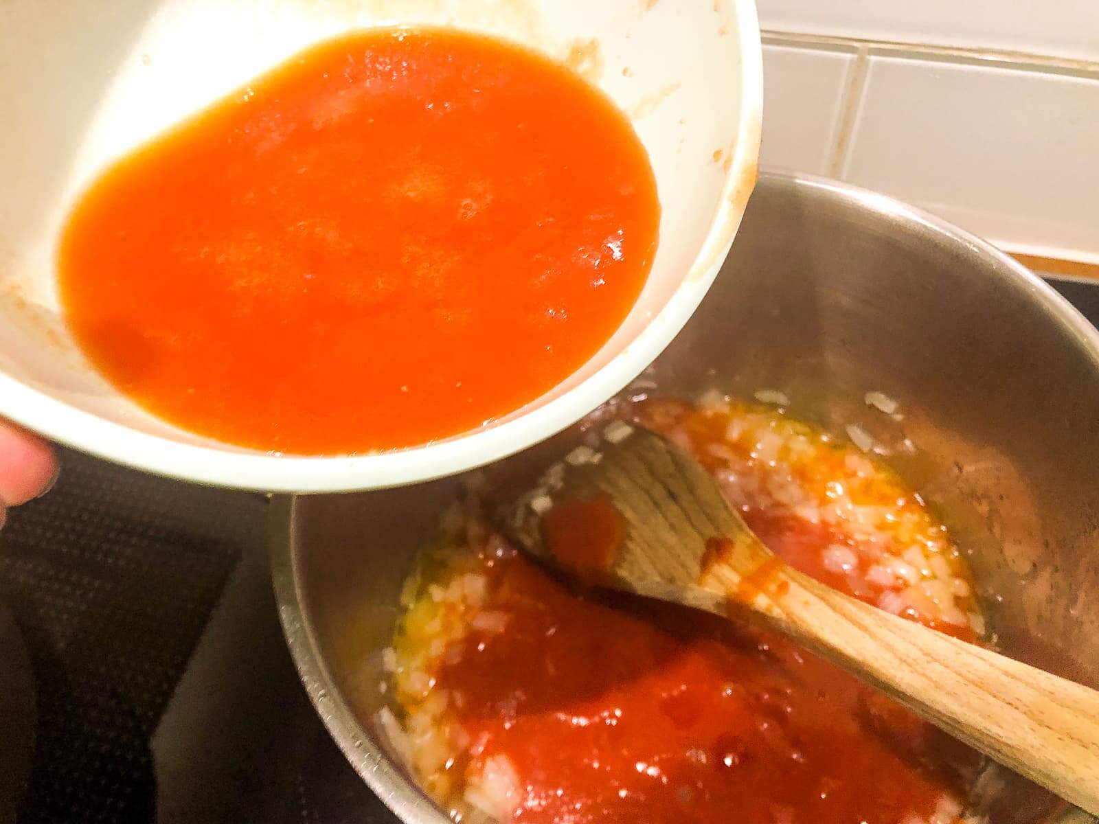 A hand adding fresh tomato juice to a pan with simmering onions and garlic.