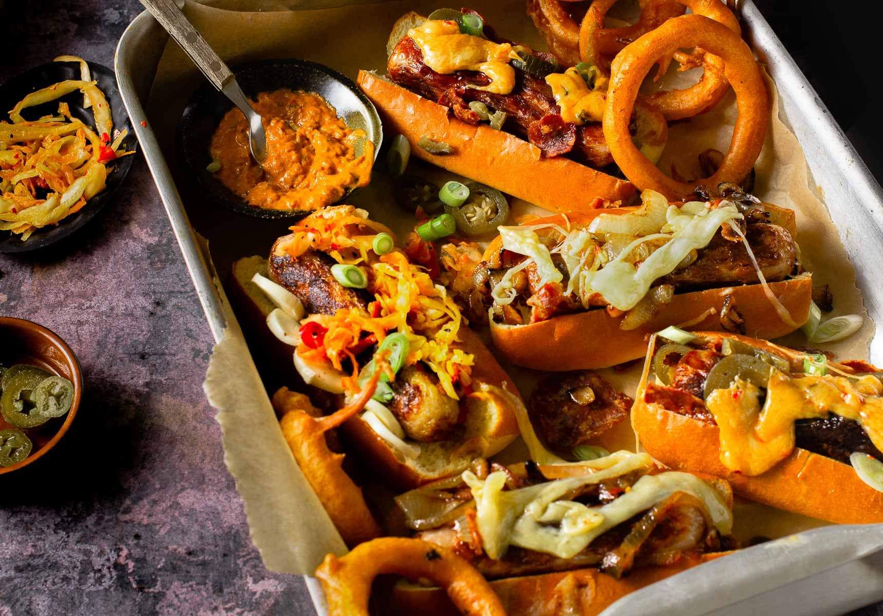 A tray of hot dogs loaded with various toppings, cheese, bacon and onions with some chillies and mustard to the side.