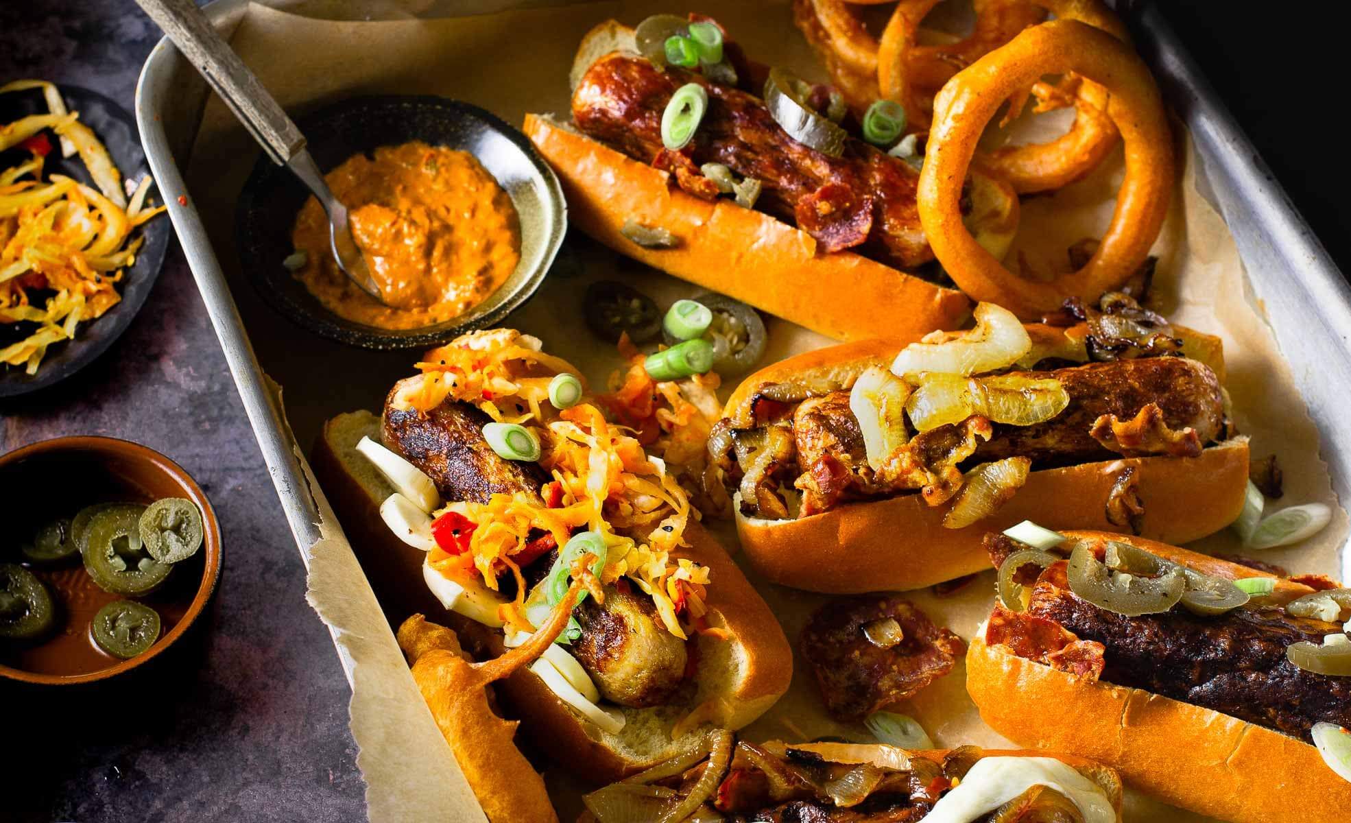 A tray containing cooked hot dogs in brioche buns with a variety of different toppings and some onion rings.