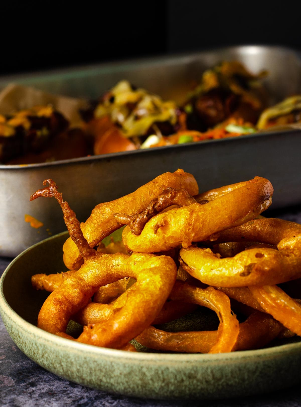 A bowl of onion rings and a full tray of loaded hot dogs to the back.