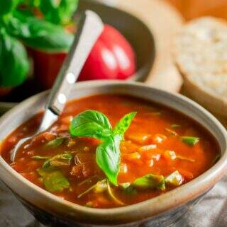 A bowl of fresh tomato & basil soup served with crusty toasted bread, topped with basil leaves and a wooden handle spoon inside the rustic bowl and fresh tomatoes to the back.