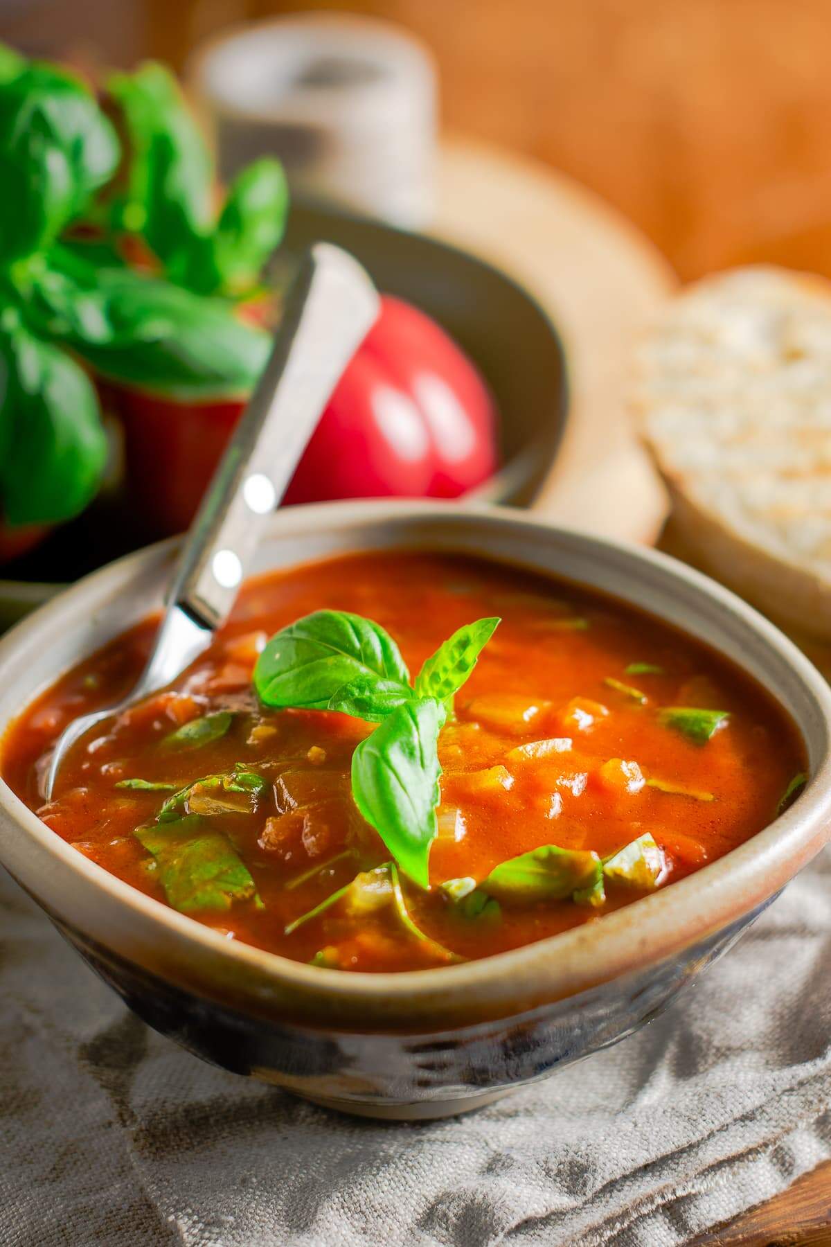 A bowl of fresh tomato & basil soup served with crusty toasted bread, topped with basil leaves and a wooden handle spoon inside the rustic bowl and fresh tomatoes to the back.