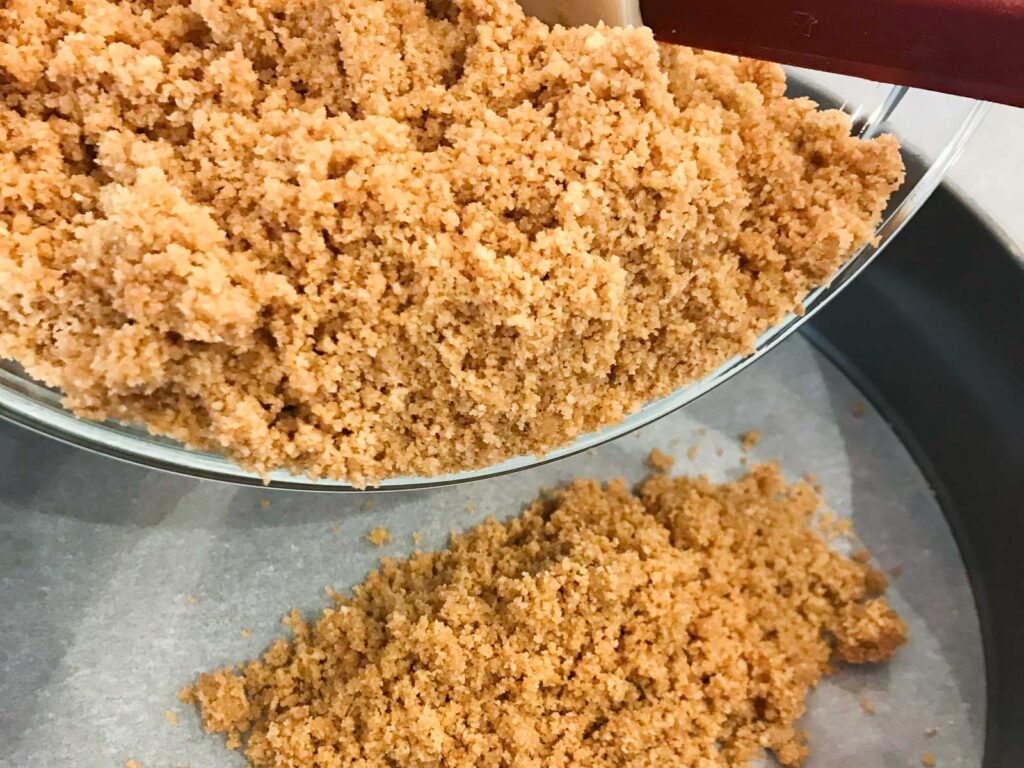 Preparing the base of a cheesecake b y adding crushed digestive biscuits and melted butter to a parchment lined springform pan.