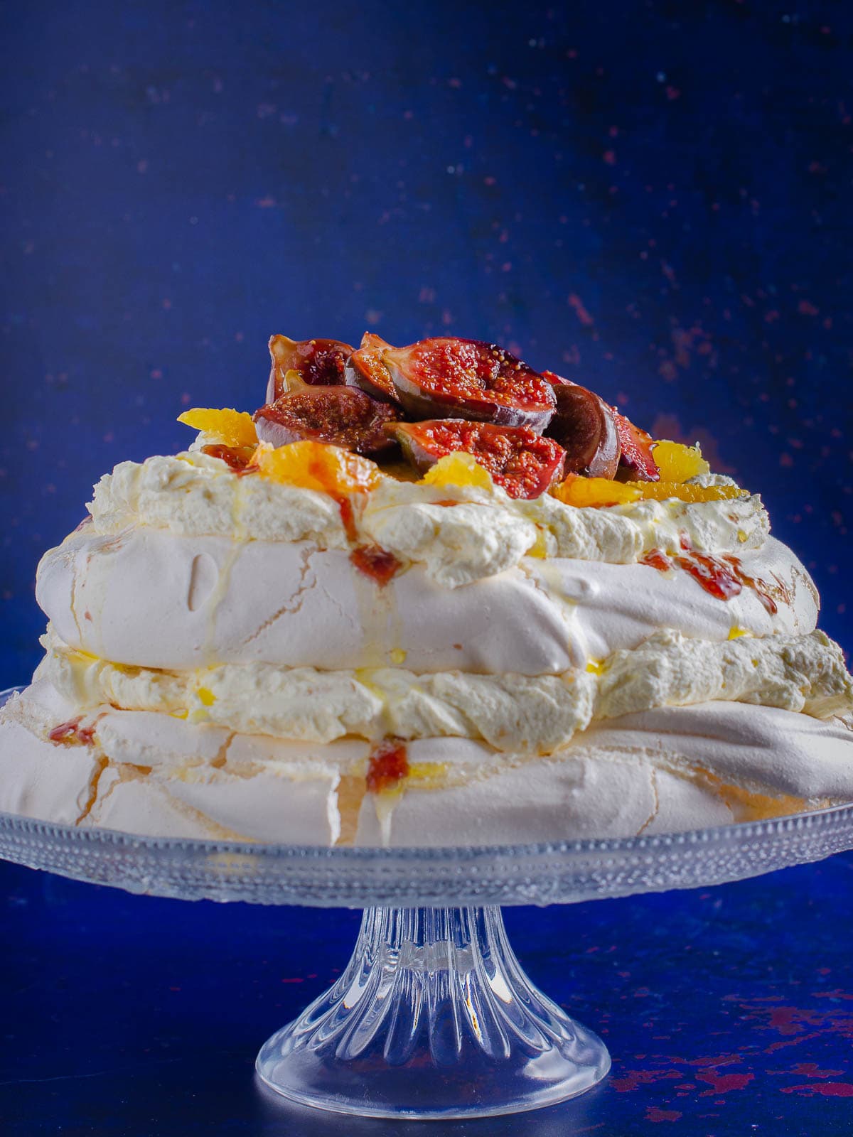 A stunning 2 layer pavlova filled with ginger cream, topped with sticky figs and orange segments served on a glass cake platter in a bright blue backdrop.