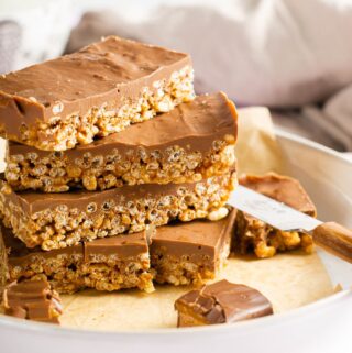 A white plate stacked with slices of Mars Bar Krispie bites topped with chocolate, a knife on the edge and some pieces of Mars bars sliced into small pieces.