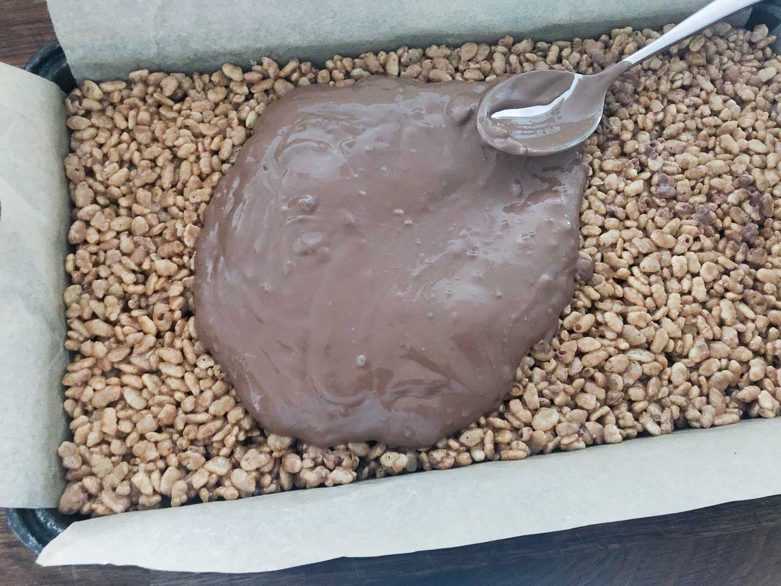 Melted chocolate being spread over an oblong pan filled with mars bar rice krispie treats lined with baking paper.