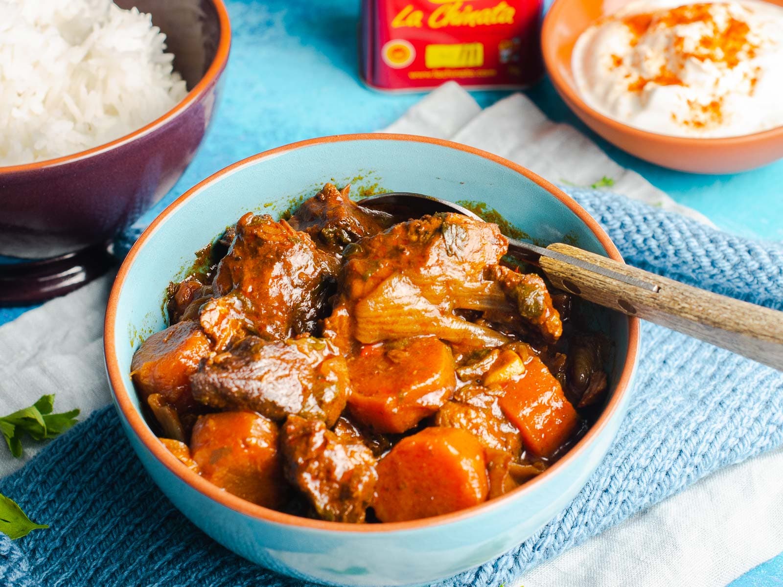 A table setting of beef paprika goulash in a blue bowl, a bowl of white rice to the back served with sour cream and a tin of La Chinata smoked paprika to the side.