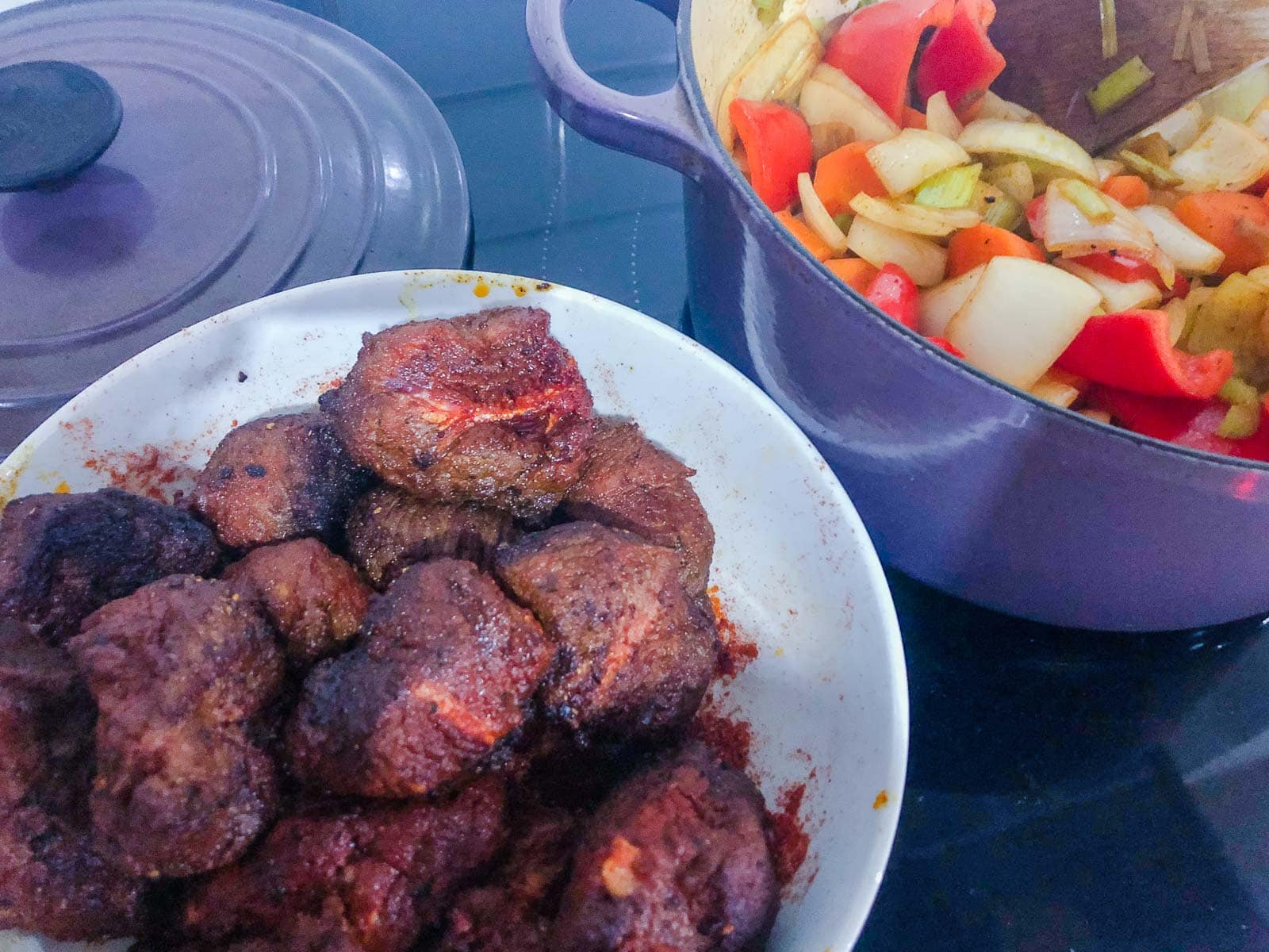 Diced beef, coated in paprika that has been browned off in a pan alongside a pan with vegetables cooking down to put together to make a beef goulash.